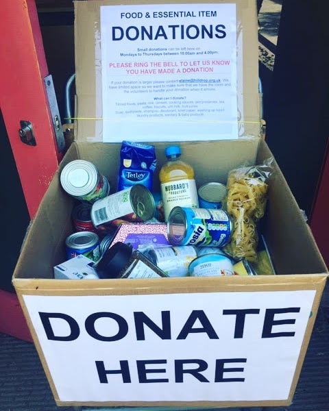 Photo of box full of food items, tins, packets of pasta, tea, cooking oil with a sign on it that reads "Donate here" and " Food & essential item donations"